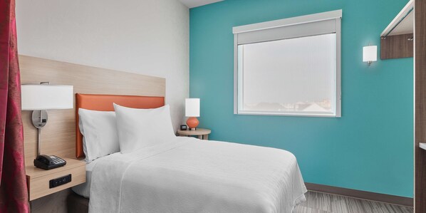 Una habitación de hotel con cama, mesita de noche, teléfono, lámpara y ventana con persianas.