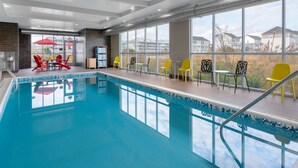Una piscina con borde de azulejos, rodeada de sillas amarillas y rojas, y grandes ventanales que ofrecen vistas a edificios.