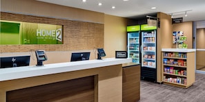 Zona de recepción de un hotel con un quiosco de registro digital, un refrigerador con aperitivos y un cartel para los ascensores.