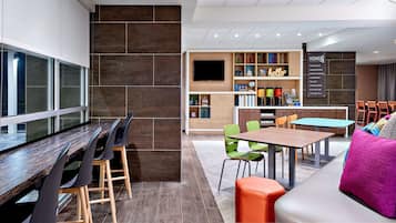 Un moderno vestíbulo de hotel con una larga mesa de comedor, sillas coloridas, un sofá con cojines vibrantes y una estantería llena de libros y objetos decorativos.