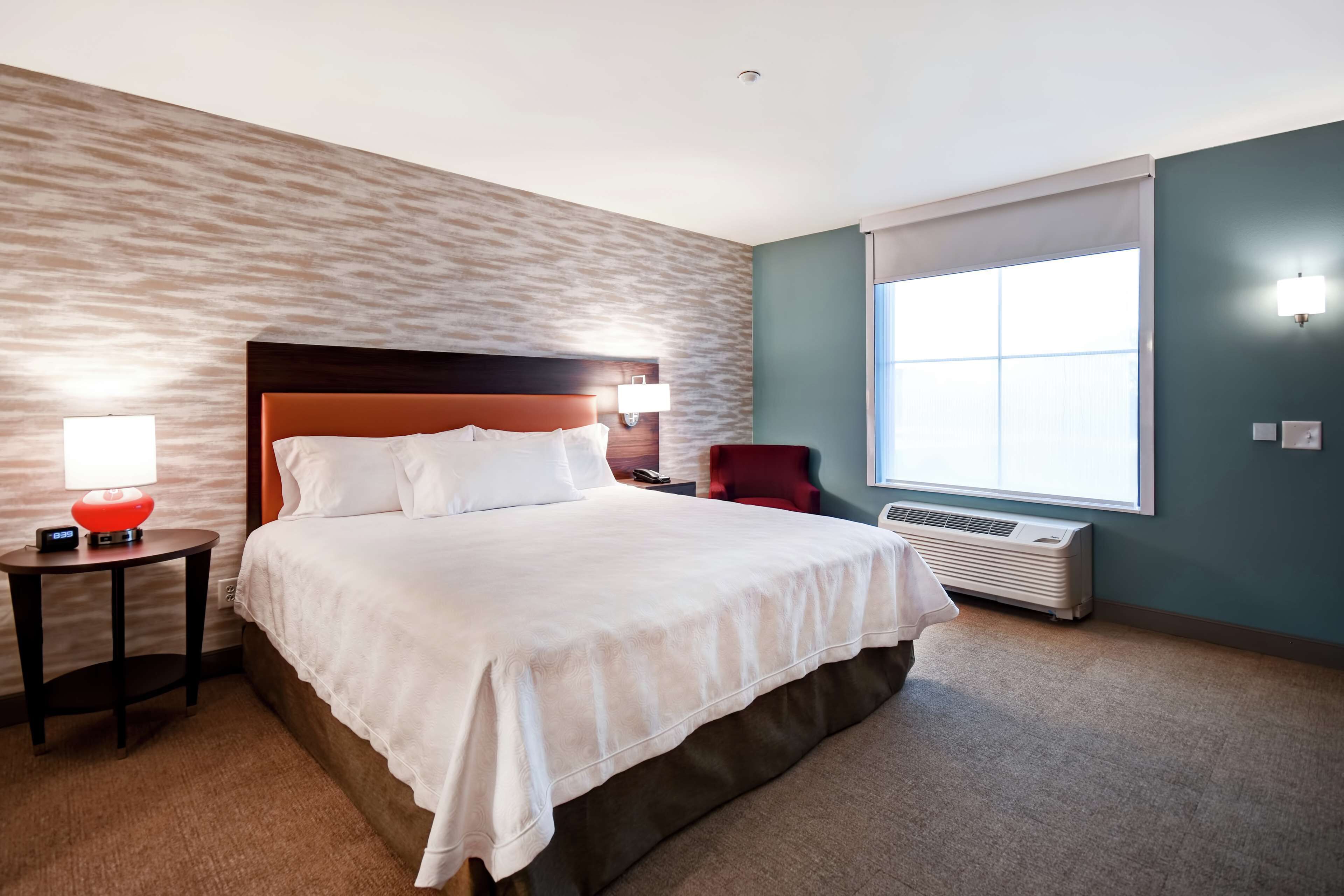 A hotel room with a large bed, a bedside table with a lamp, a chair, a window with blinds, and an air conditioning unit.