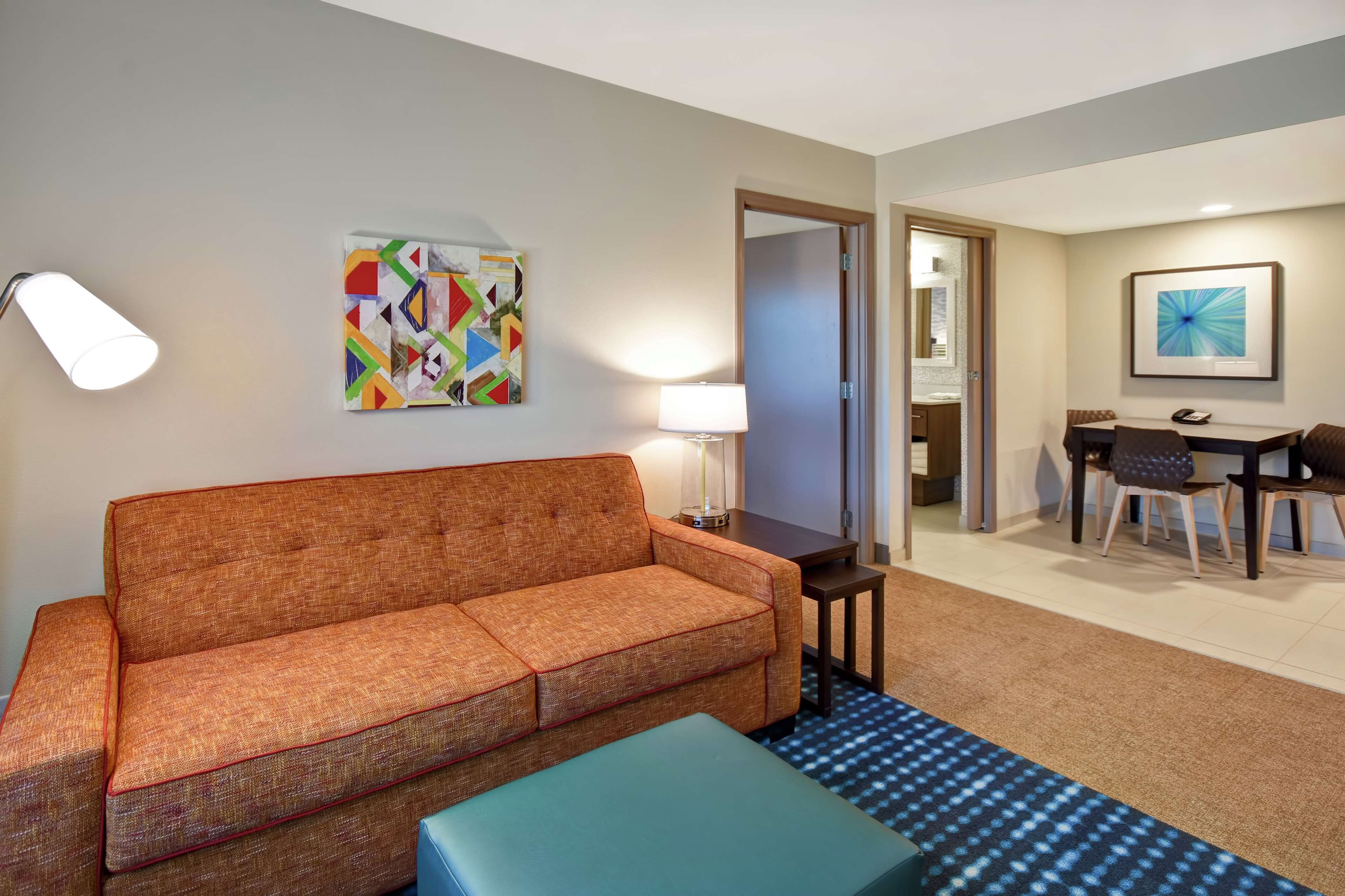 Une chambre d’hôtel avec un canapé, une petite table avec une lampe, un coin repas avec des chaises et un tableau au mur.