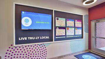 A hotel lobby with a digital screen displaying 'WELCOME TO TRU' and 'LIVE TRU-LY LOCAL'. There are bulletin boards with various notes and a purple polka dot wall design.