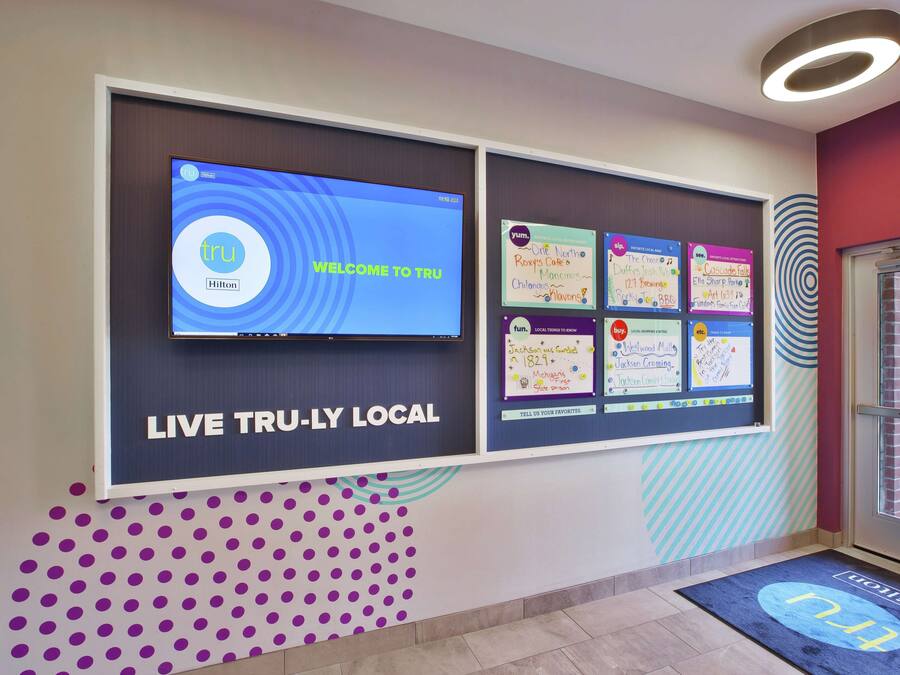 A hotel lobby with a digital screen displaying 'WELCOME TO TRU' and 'LIVE TRU-LY LOCAL'. There are bulletin boards with various notes and a purple polka dot wall design.