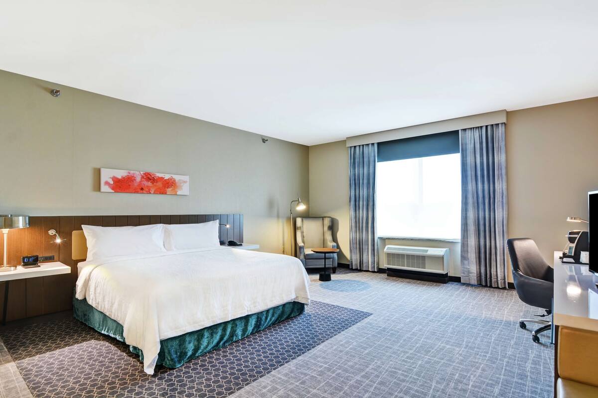 A hotel room with a large bed, a desk, a chair, a desk lamp, a wall-mounted TV, and a window with curtains.
