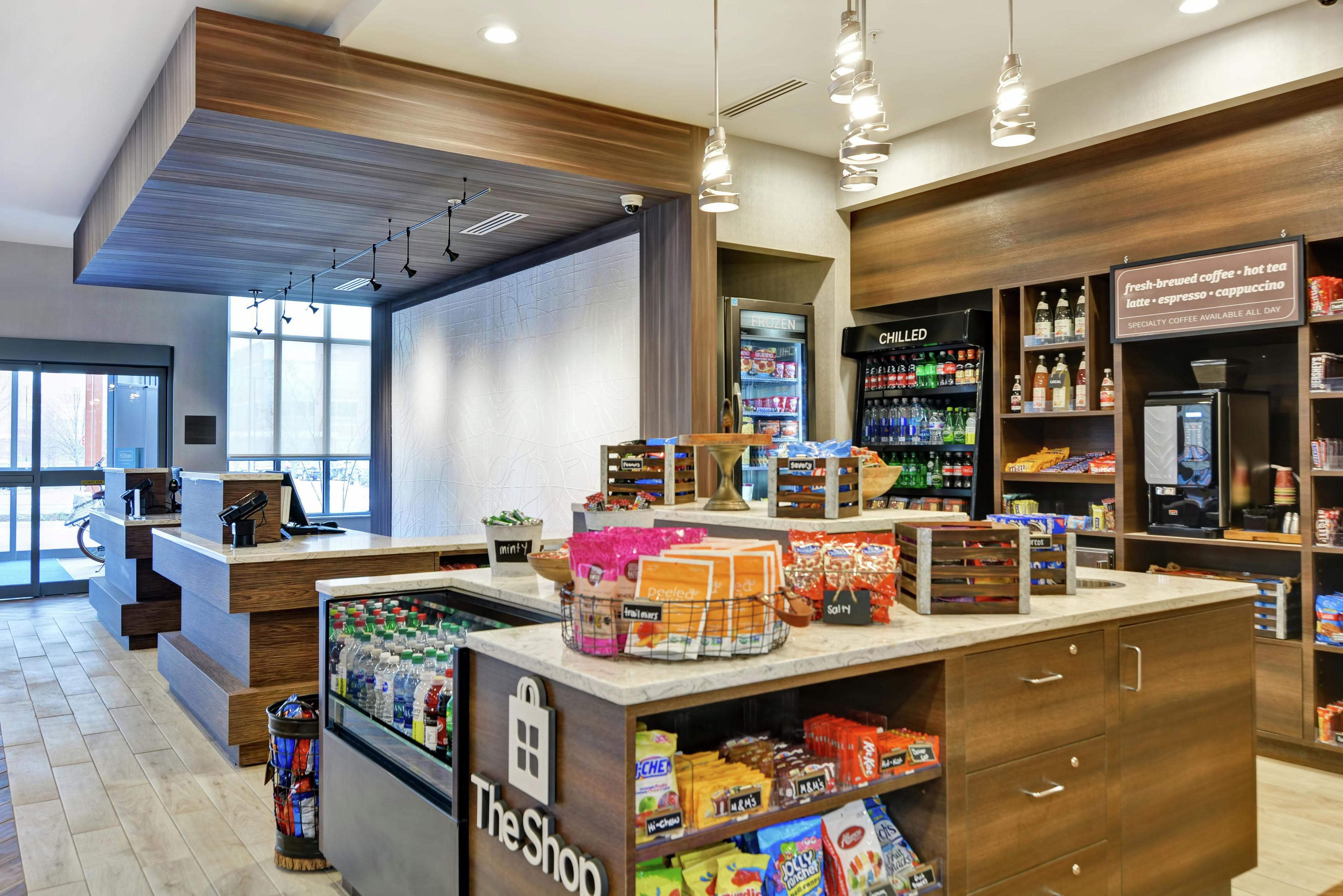 Interior de una cafetería con mostrador, estantes llenos de aperitivos y bebidas, y un refrigerador con productos frescos.