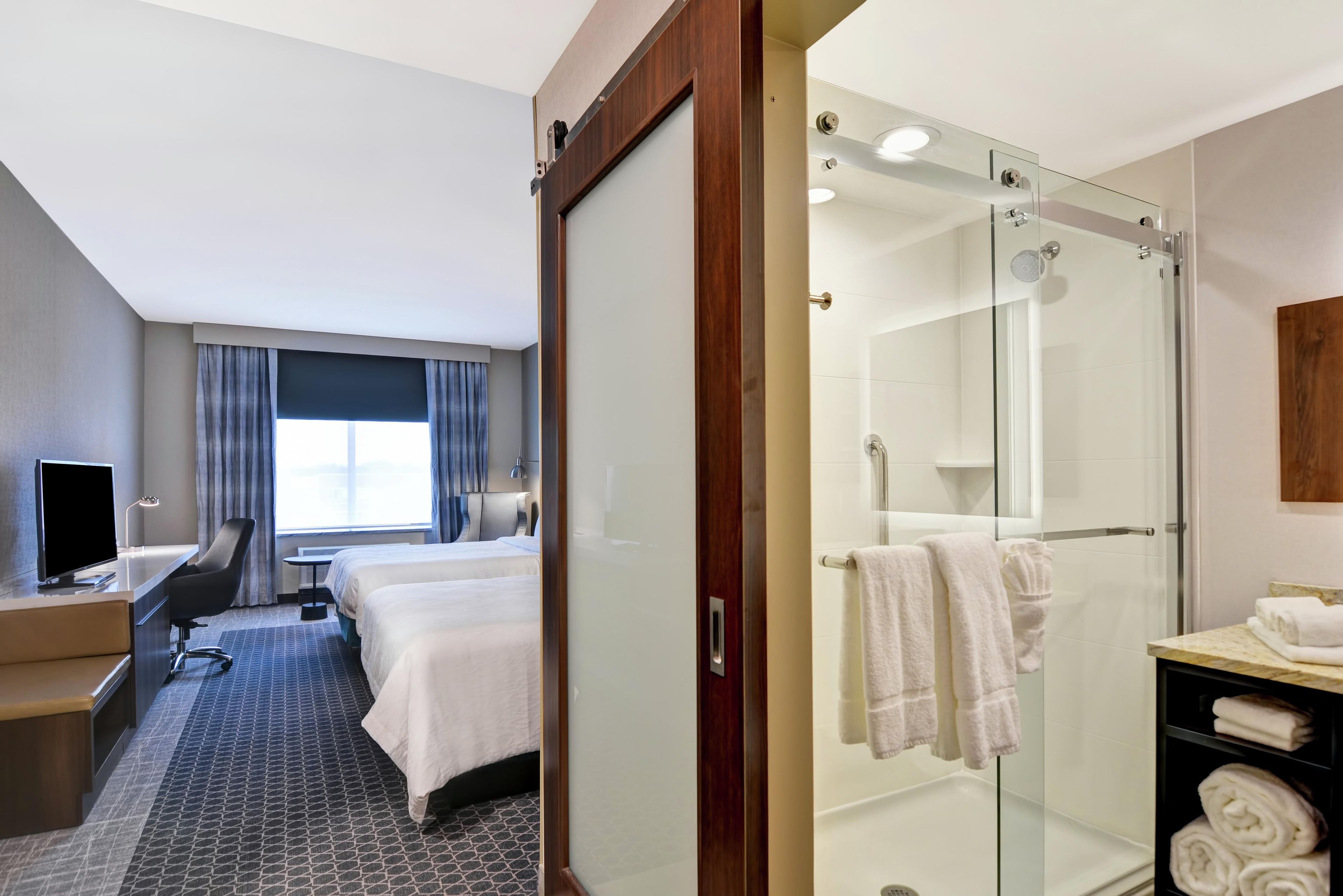 A hotel room with a bed, desk, chair, TV, and a shower area with towels.