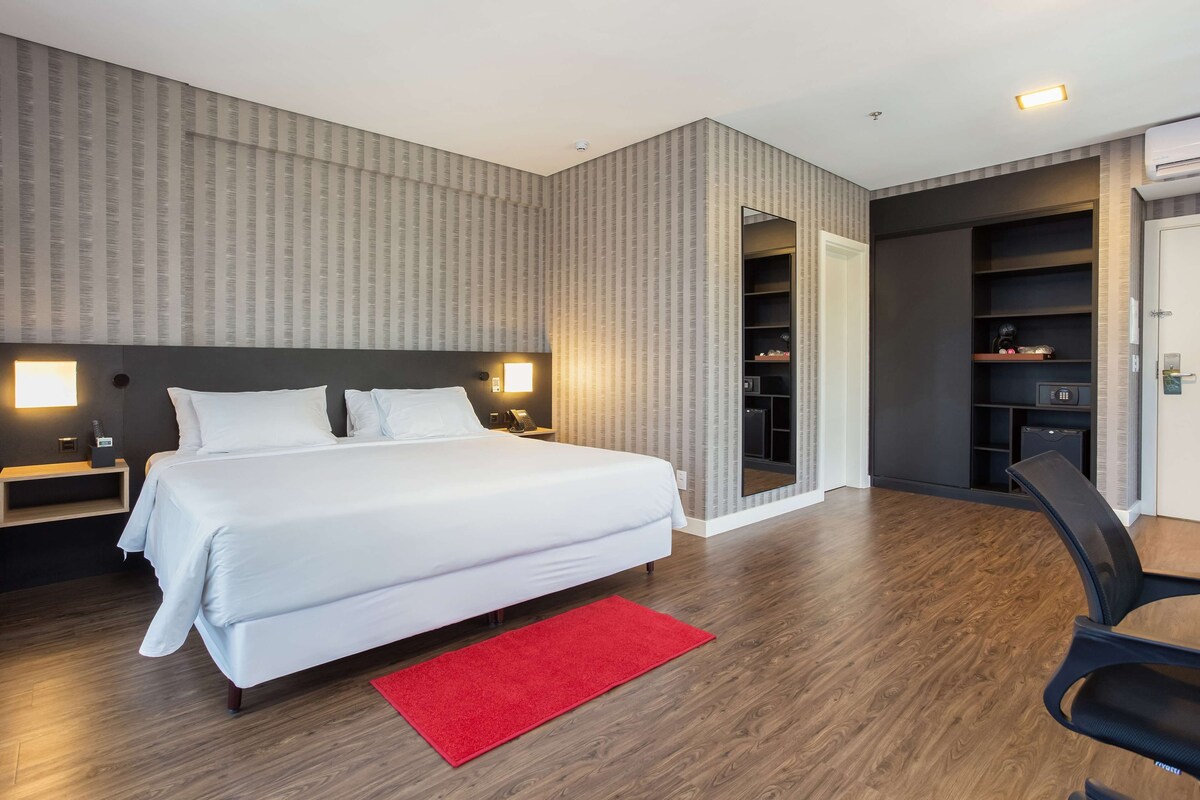 A modern hotel room with a large bed, bedside tables, a red rug, a desk, and a built-in wardrobe.