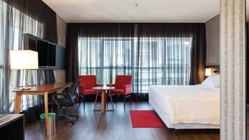 A hotel room with a bed, a desk, two red chairs, a TV, and large windows with curtains.