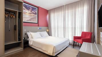 Ein Hotelzimmer mit einem Bett, einem roten Sessel, einem Kleiderschrank und einem großen Fenster mit Vorhängen.