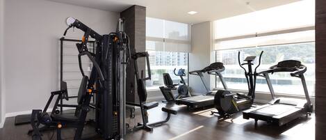 A modern gym with cardio machines, weight training equipment, and large windows offering views of the city.