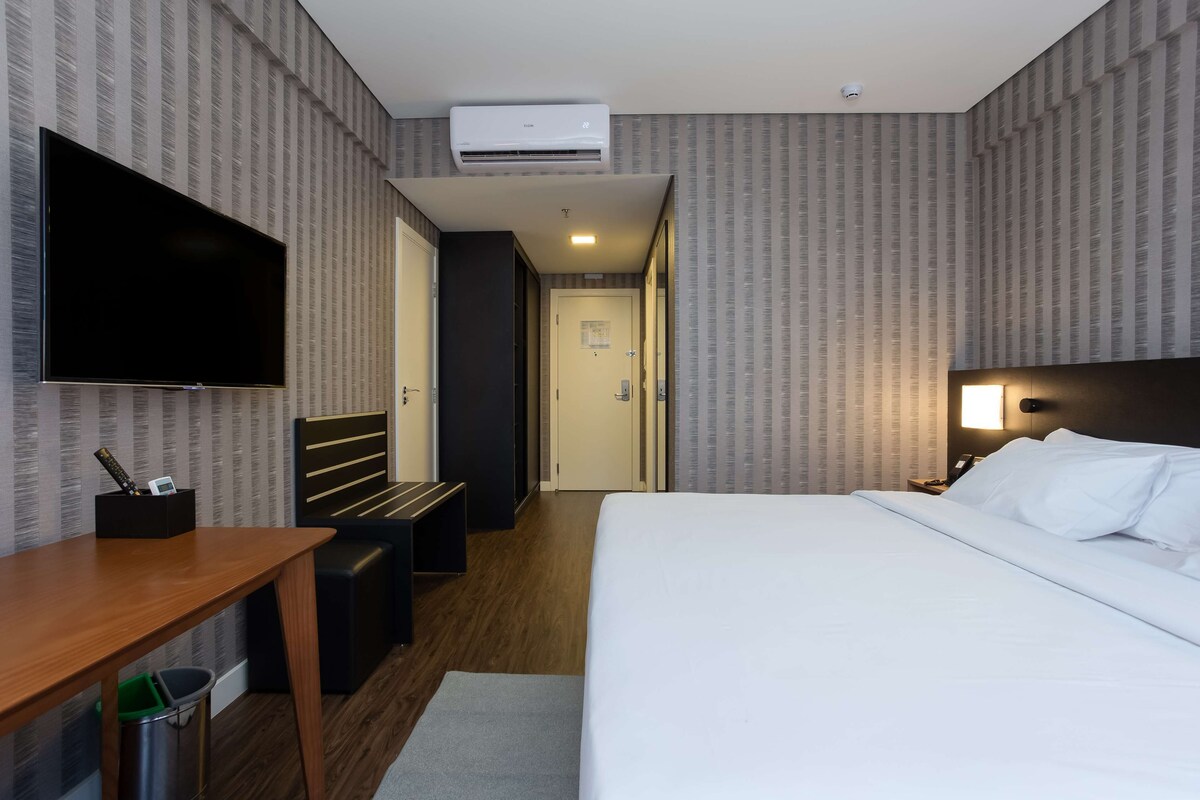 A hotel room with a bed, a desk, a TV, and an air conditioning unit.