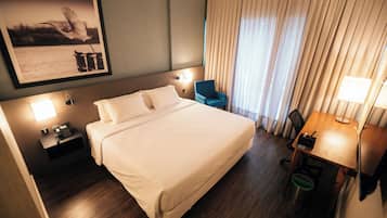 Habitación de hotel con una cama grande, lámparas de noche, un escritorio con silla, un cuadro en la pared y una ventana con cortinas.