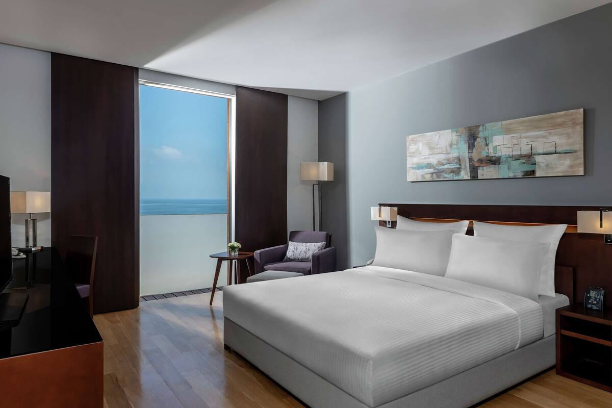 A modern hotel room with a large bed, a desk with a flat-screen TV, a chair, a small table with a lamp, and a view of the sea through a large window.