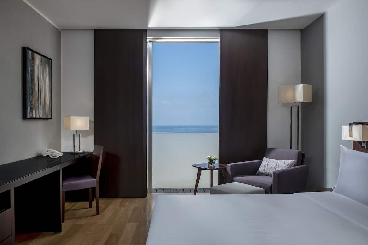 A hotel room with a bed, a desk with a phone, a chair, a small table with a vase, and a lamp.