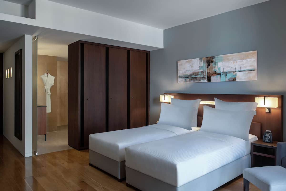 A modern hotel room with two single beds, a wooden wardrobe, a painting on the wall, and a bedside table with a lamp.