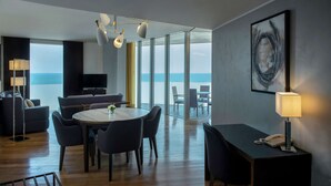 A modern hotel room with a large window offering a view of the ocean, a round dining table, a sofa, a TV, and a desk with a lamp. - Hilton Beirut Downtown (Beirut)
