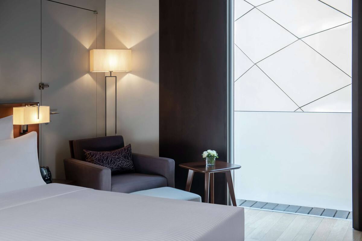 A modern hotel room with a large bed, a sitting area with a chair, a small table with a vase of flowers, and a large window with geometric patterns.