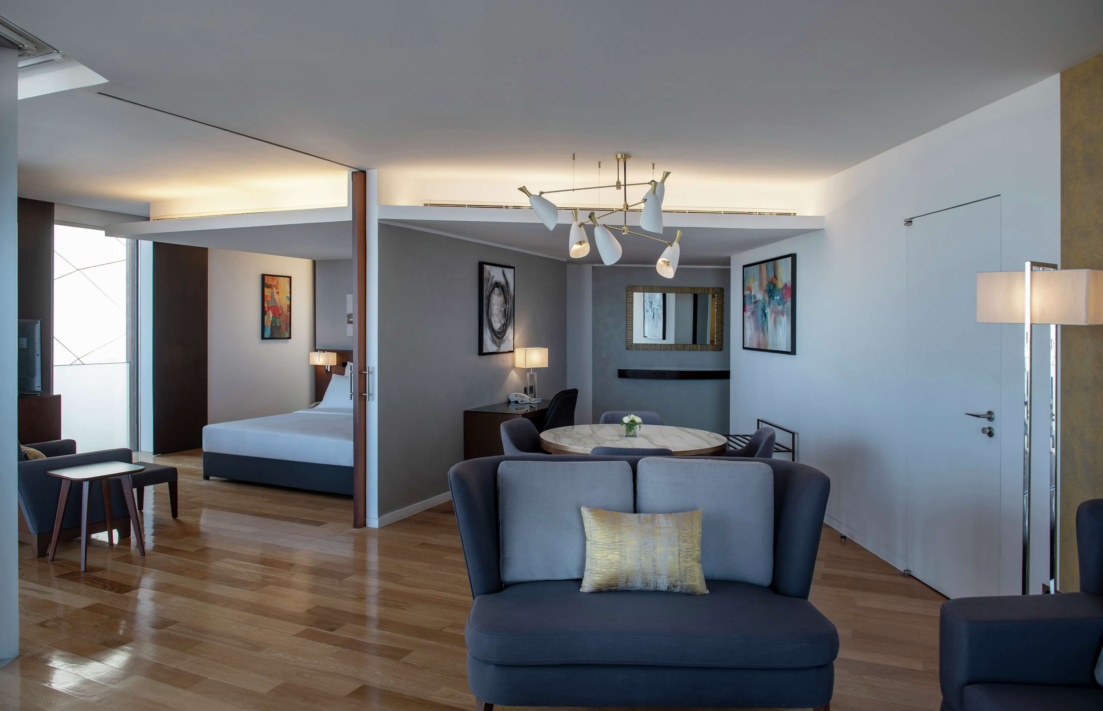 A modern hotel room with a large bed, a seating area with a sofa and armchairs, a dining table, and a chandelier.