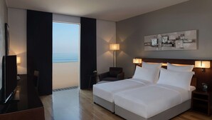 A hotel room with a large bed, two bedside tables with lamps, a desk with a chair, a TV, and a view of the sea through a window. - Hilton Beirut Downtown (Beirut)