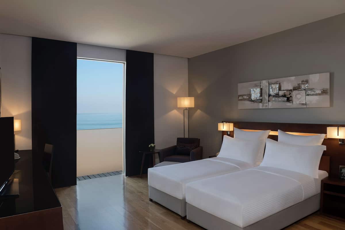 A hotel room with a large bed, two bedside tables with lamps, a desk with a chair, a TV, and a view of the sea through a window.