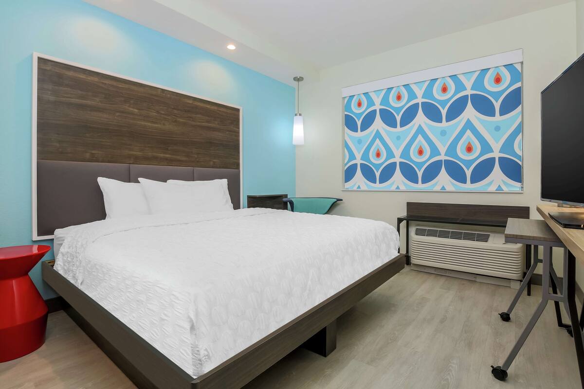 A modern hotel room with a large bed, a desk, a red chair, and a wall-mounted TV.