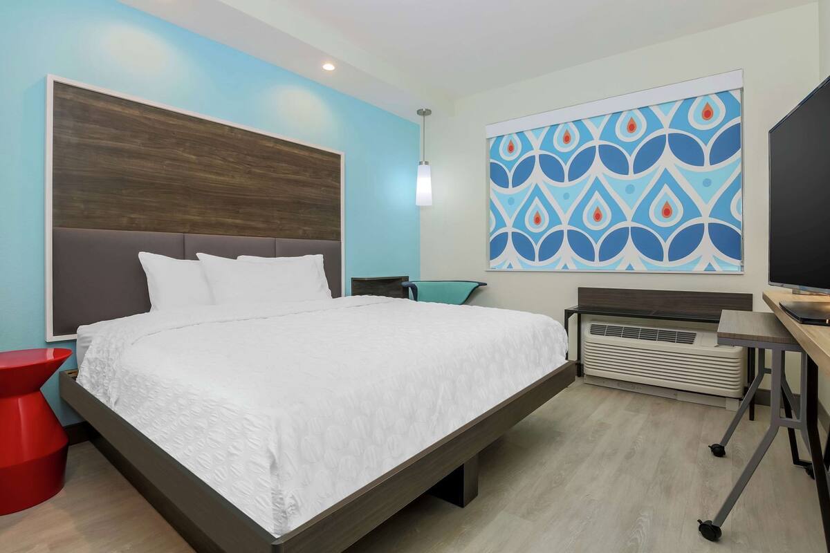 A modern hotel room with a large bed, a desk, a TV, and a wall-mounted artwork.