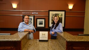 Two receptionists at a hotel counter with framed pictures on the wall and a 'Welcome to Gallup' sign. - Hampton Inn & Suites Gallup (Gallup)