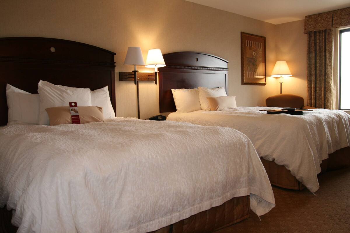 Ein Hotelzimmer mit zwei Betten, jeweils mit weißer Bettwäsche und einem Holzkopfstück. Es gibt zwei Nachttischlampen und einen Nachttisch mit einer Lampe. Das Zimmer hat ein großes Fenster mit Vorhängen und ein gerahmtes Bild an der Wand.
