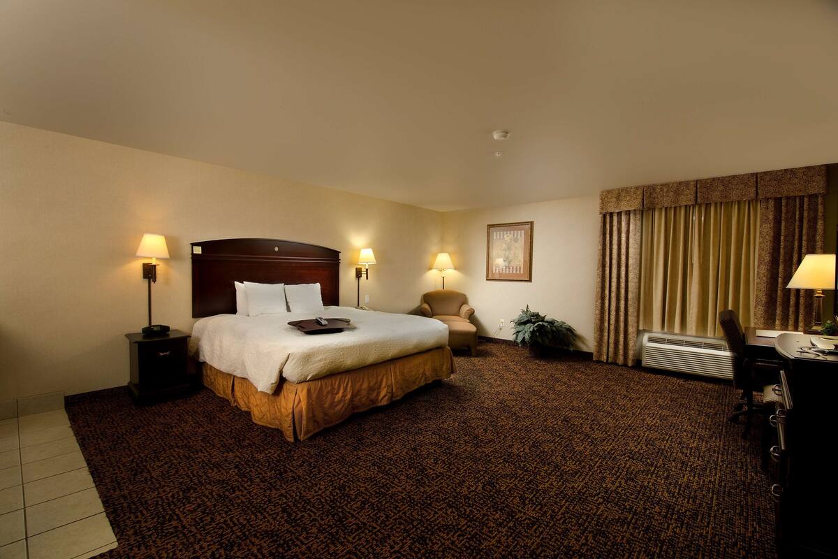 Ein Hotelzimmer mit einem großen Bett, zwei Nachttischlampen, einem Schreibtisch mit Stuhl, einer Zimmerpflanze und einem fenster mit Vorhängen.