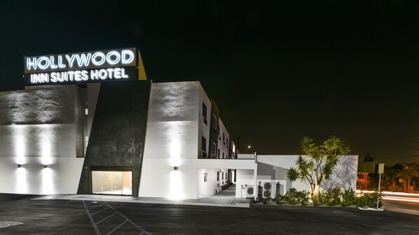 The exterior of the Hollywood Inn Suites Hotel with a prominent neon sign, a modern architectural design, and a parking lot. - Hollywood Inn Suites Hotel (Gardena)