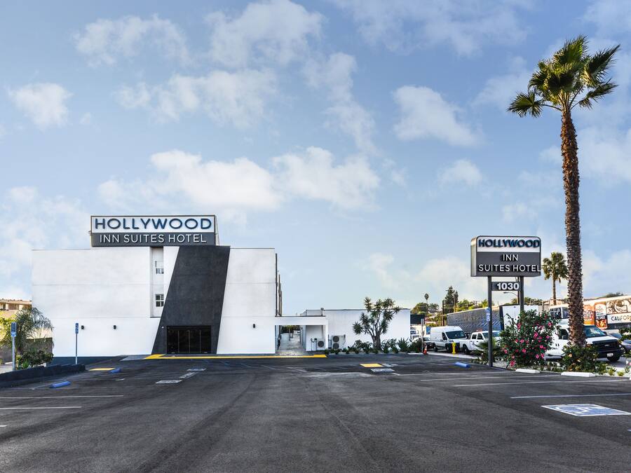 A modern building with a sign reading "Hollywood Inn Suites Hotel" and a parking lot in front.