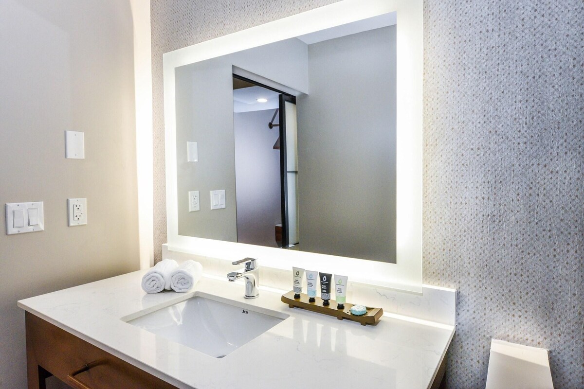 A modern bathroom with a large mirror, white sink, and two towel rolls.
