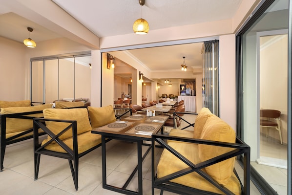 Un espace repas avec des tables et des chaises, une cuisine à l’arrière-plan et une porte-fenêtre coulissante.