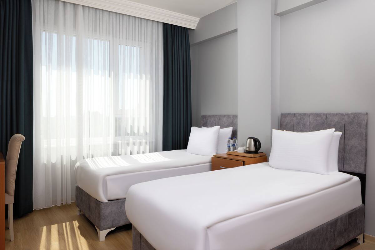 Ein Hotelzimmer mit zwei Betten, einem Schreibtisch, einem Stuhl, einem Nachttisch mit Lampe und einem Fenster mit durchsichtigen Vorhängen.