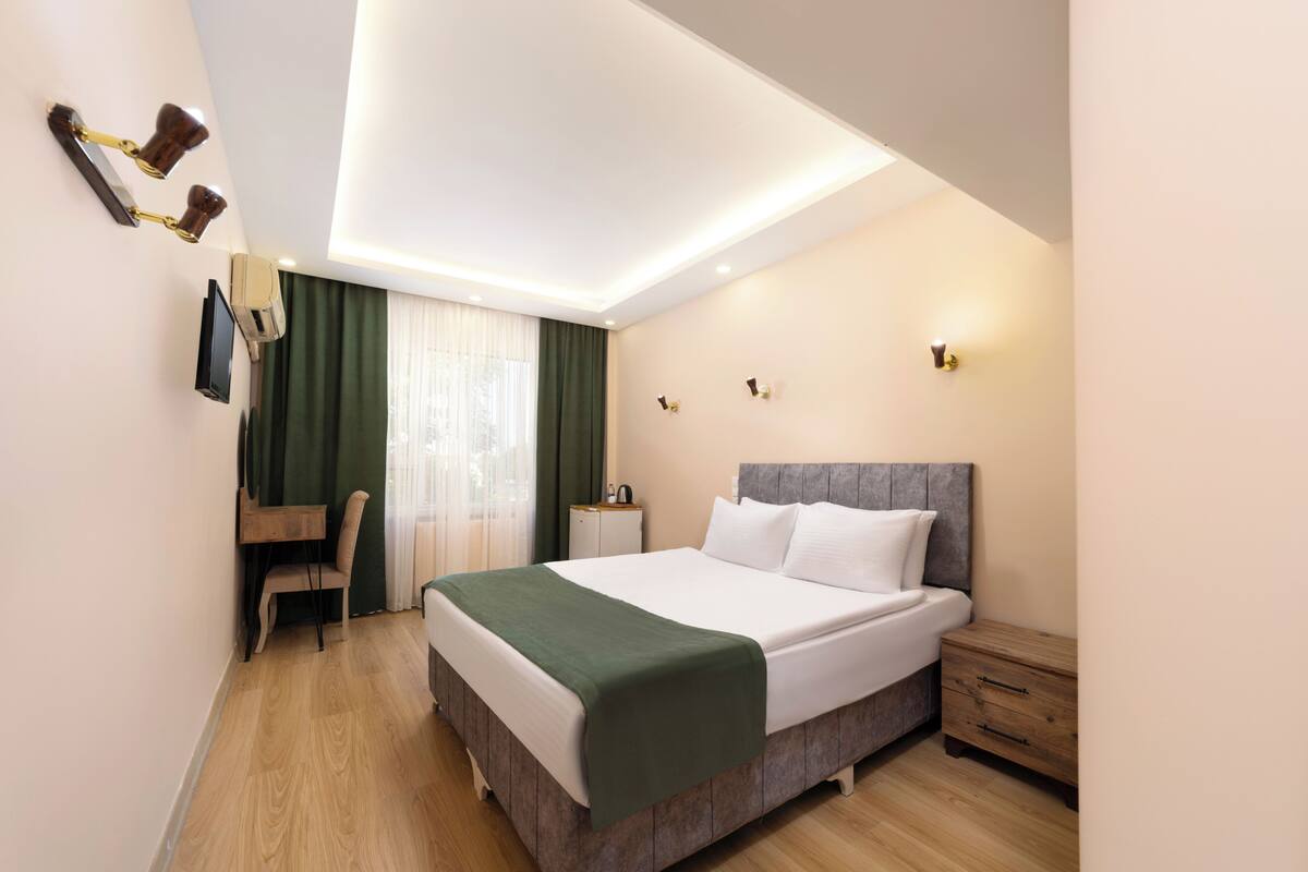 Ein Hotelzimmer mit Bett, Nachttisch, Schreibtisch, Stuhl, Fernseher und einem Fenster mit Vorhängen.