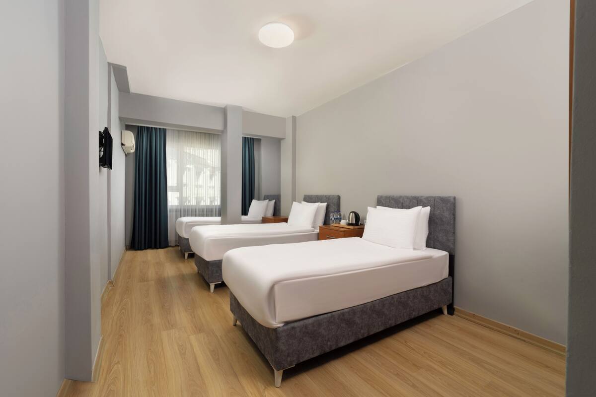 Ein Hotelzimmer mit zwei Betten, Holzboden, Deckenleuchte und einem Fenster mit Vorhängen.