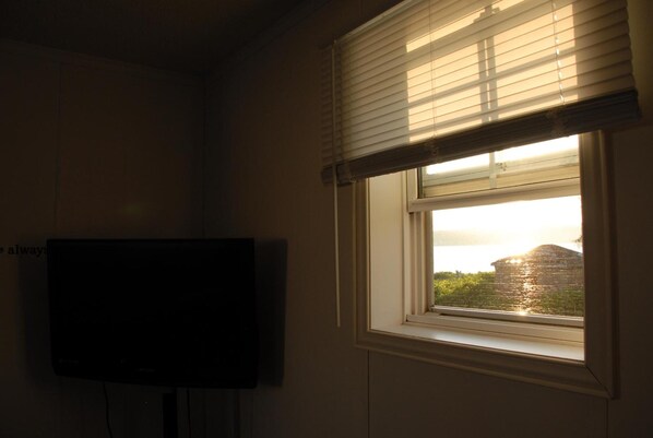 A room with a television mounted on the wall and a window with blinds, overlooking a body of water and buildings.