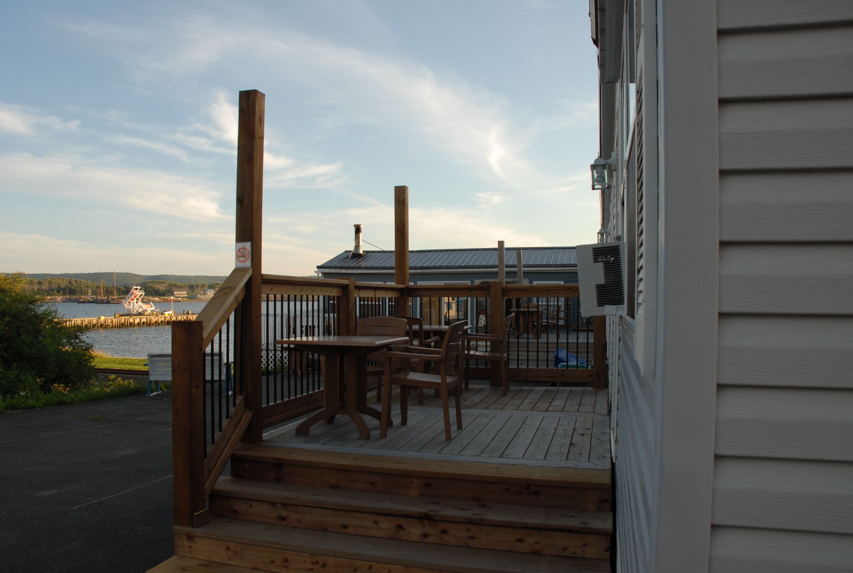 A wooden deck with outdoor seating, a view of a body of water, and a building with a metal roof in the background.