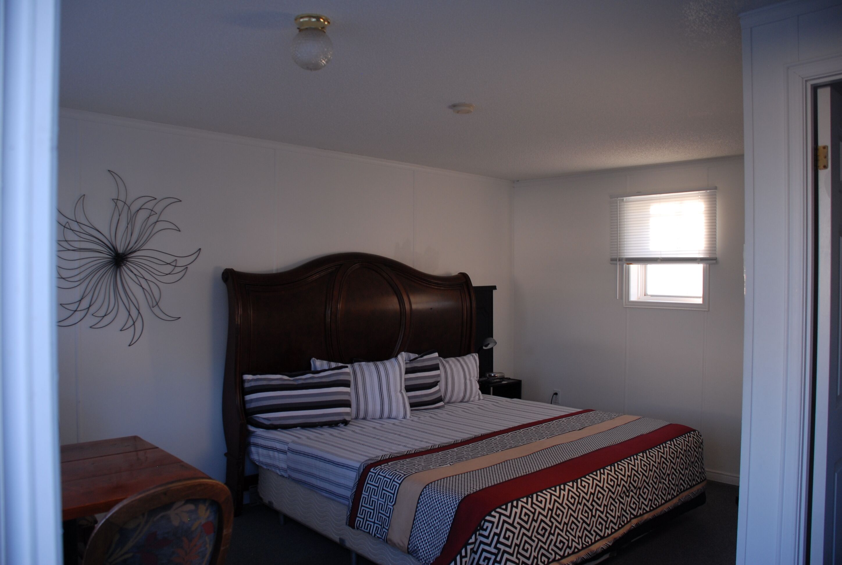 Un dormitorio con cabecera de madera, cama con ropa de cama a rayas, un adorno en la pared, una mesita de noche y una ventana con persianas.