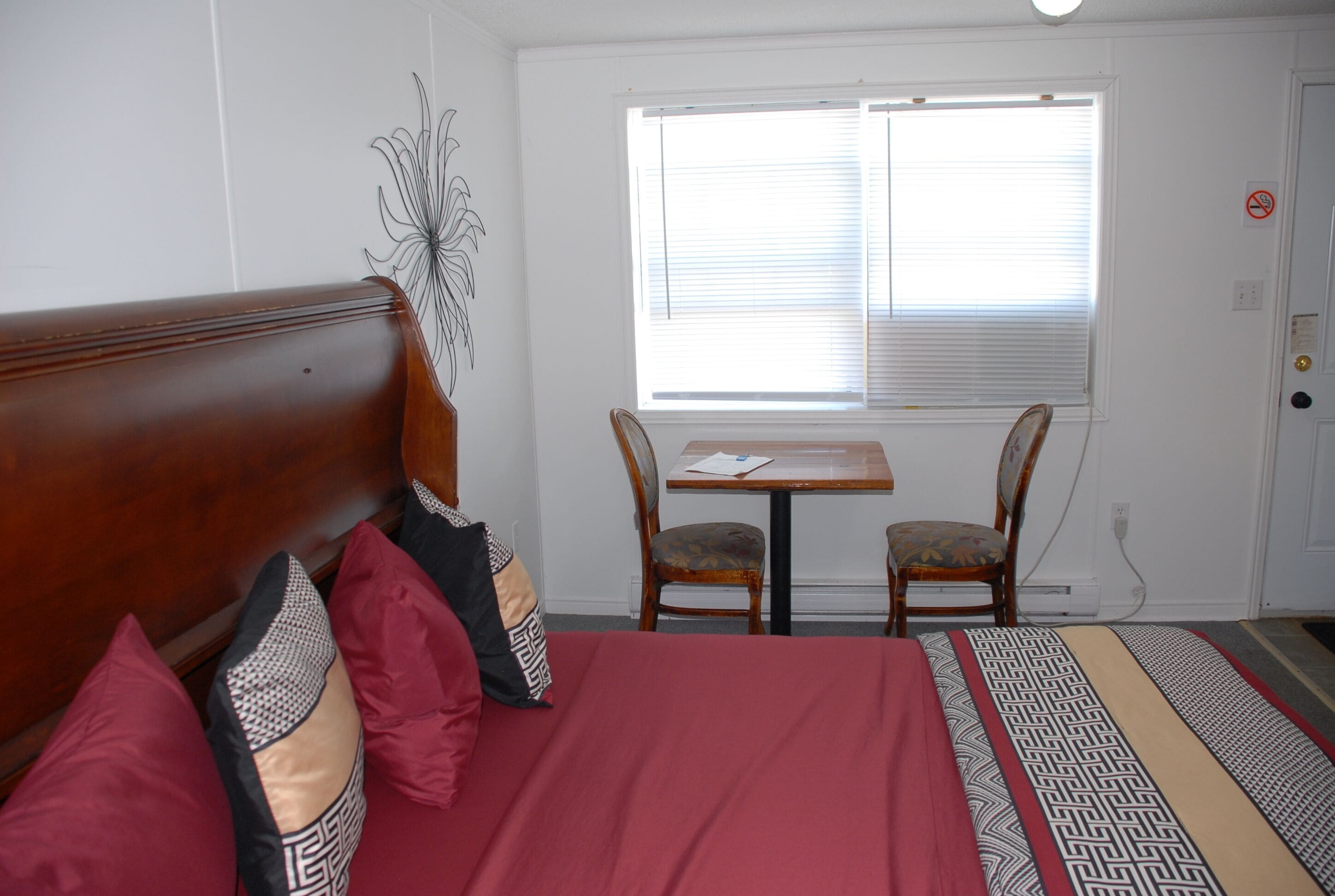 Un dormitorio con cabecera de madera, colcha roja, dos almohadas estampadas, una mesita con una silla y una ventana con persianas.