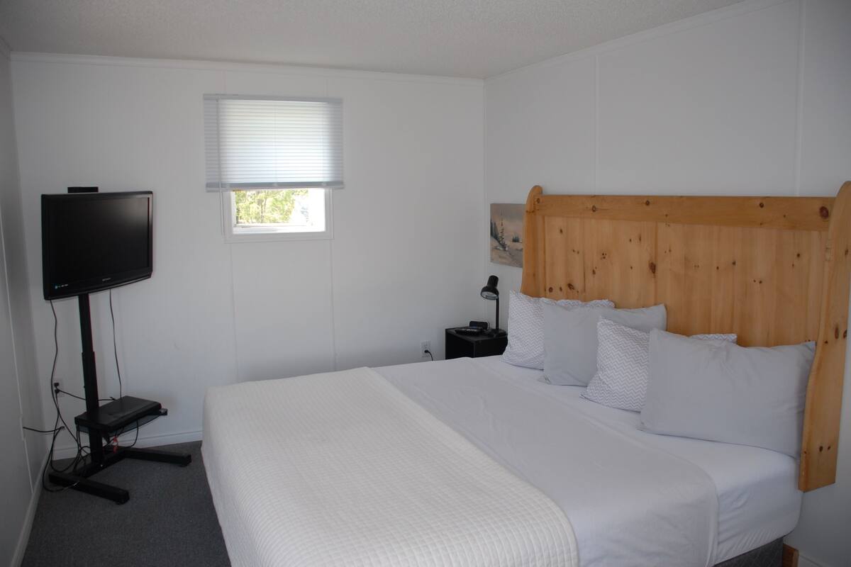 Une chambre à coucher avec un grand lit, un téléviseur posé sur un meuble, une petite table et une fenêtre avec des stores.