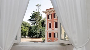 Una ventana con cortinas blancas, vista de un edificio con fachada roja y varias ventanas, y un árbol al fondo.