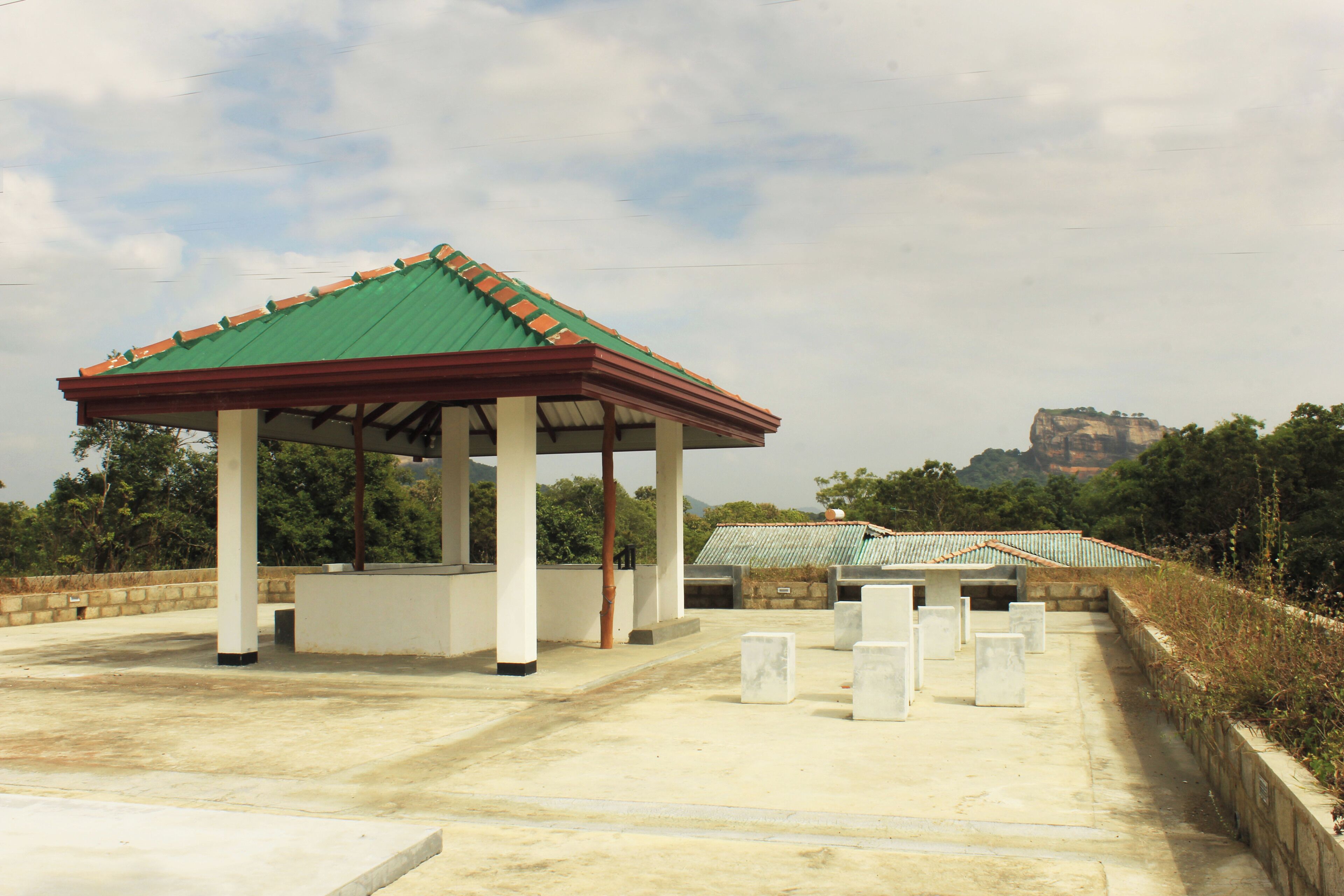 A covered pavilion with a green roof and white walls, surrounded by concrete pillars and benches.
