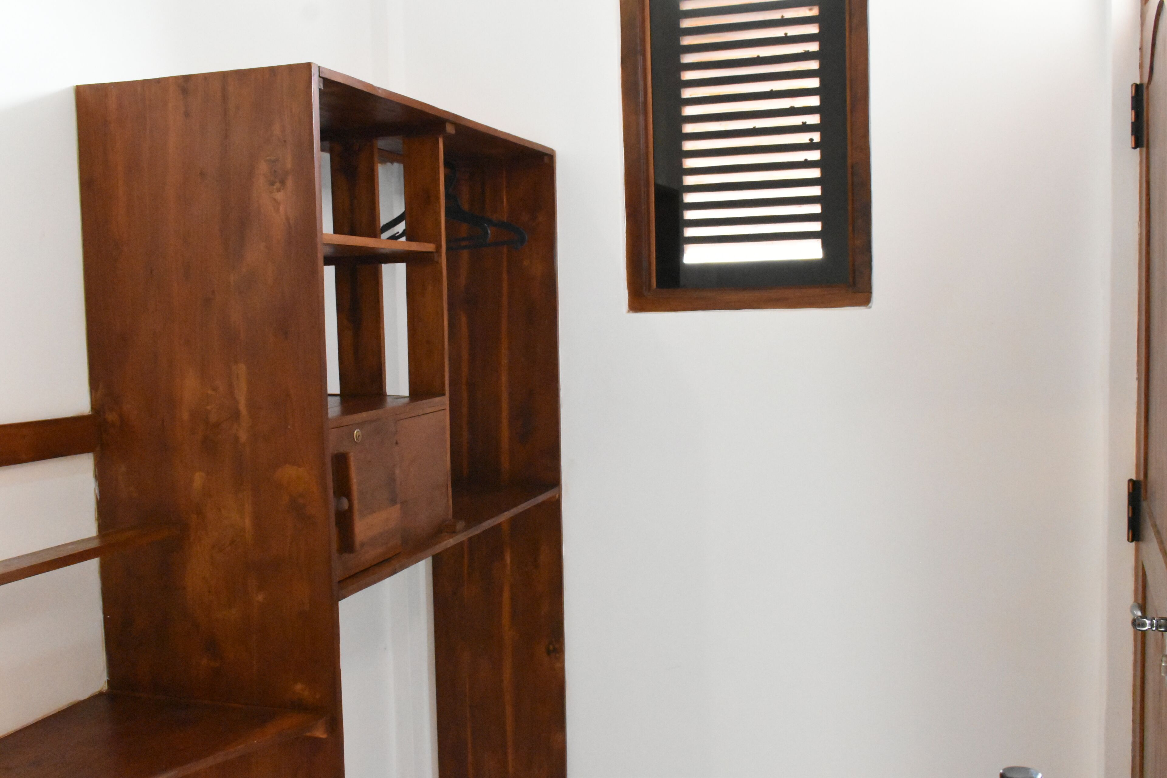 A wooden wardrobe with multiple shelves and a hanging rod, positioned next to a window with blinds.