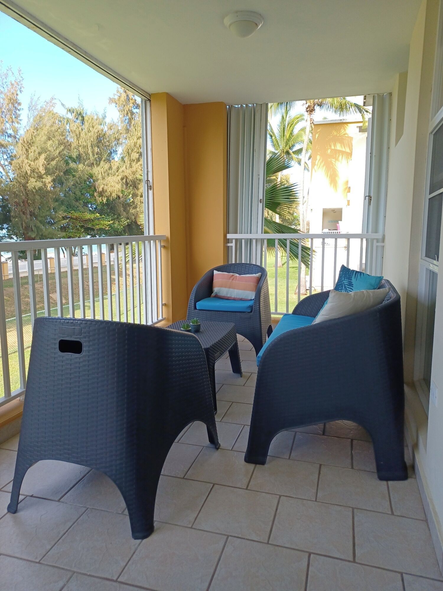 Ein Balkon mit zwei Korbstühlen, einem kleinen Tisch, einer gelben Wand und Blick auf Bäume und ein Gebäude.