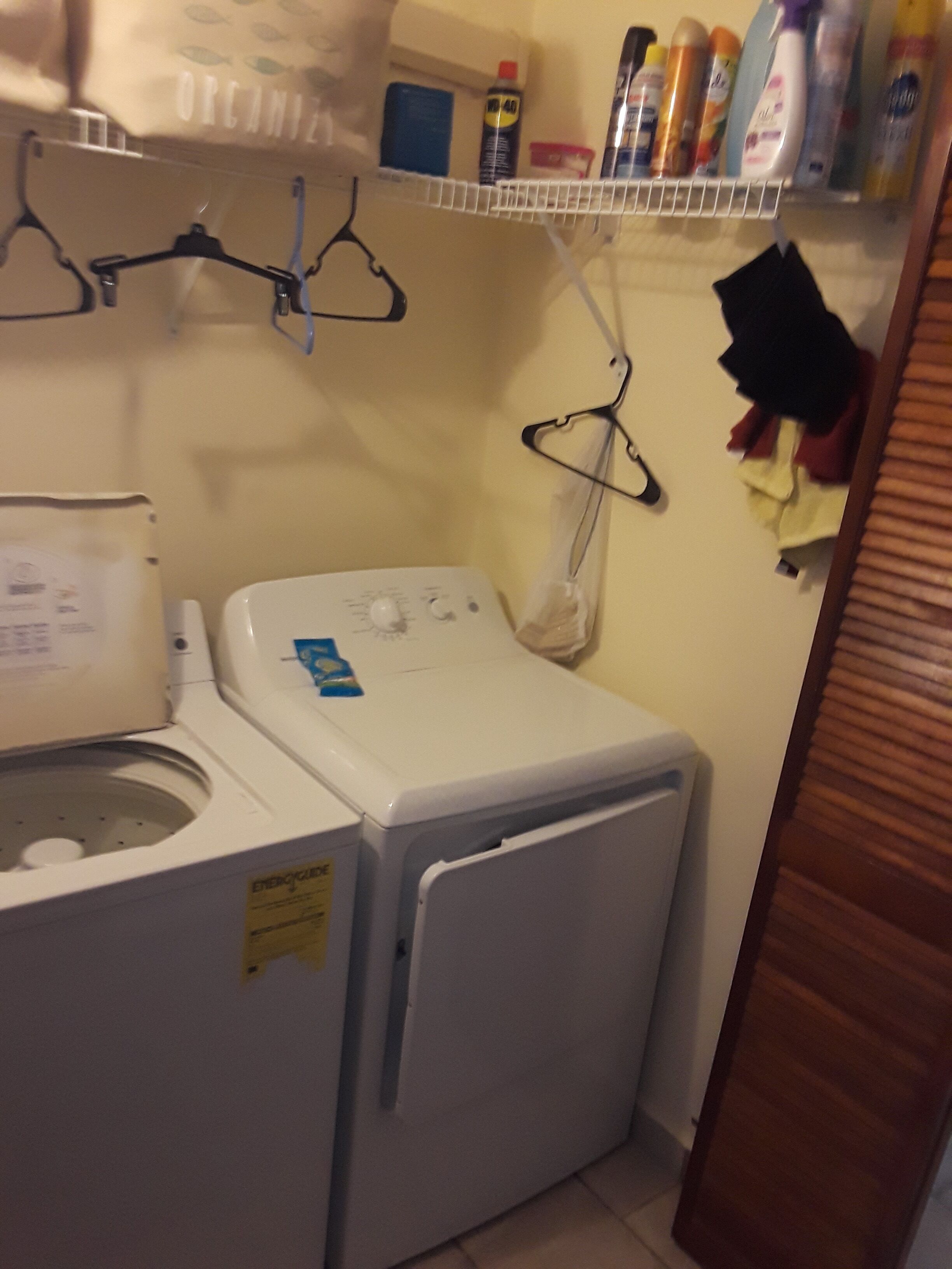 A compact laundry area with a front-loading washing machine, a drying rack, and a shelf with various cleaning products.
