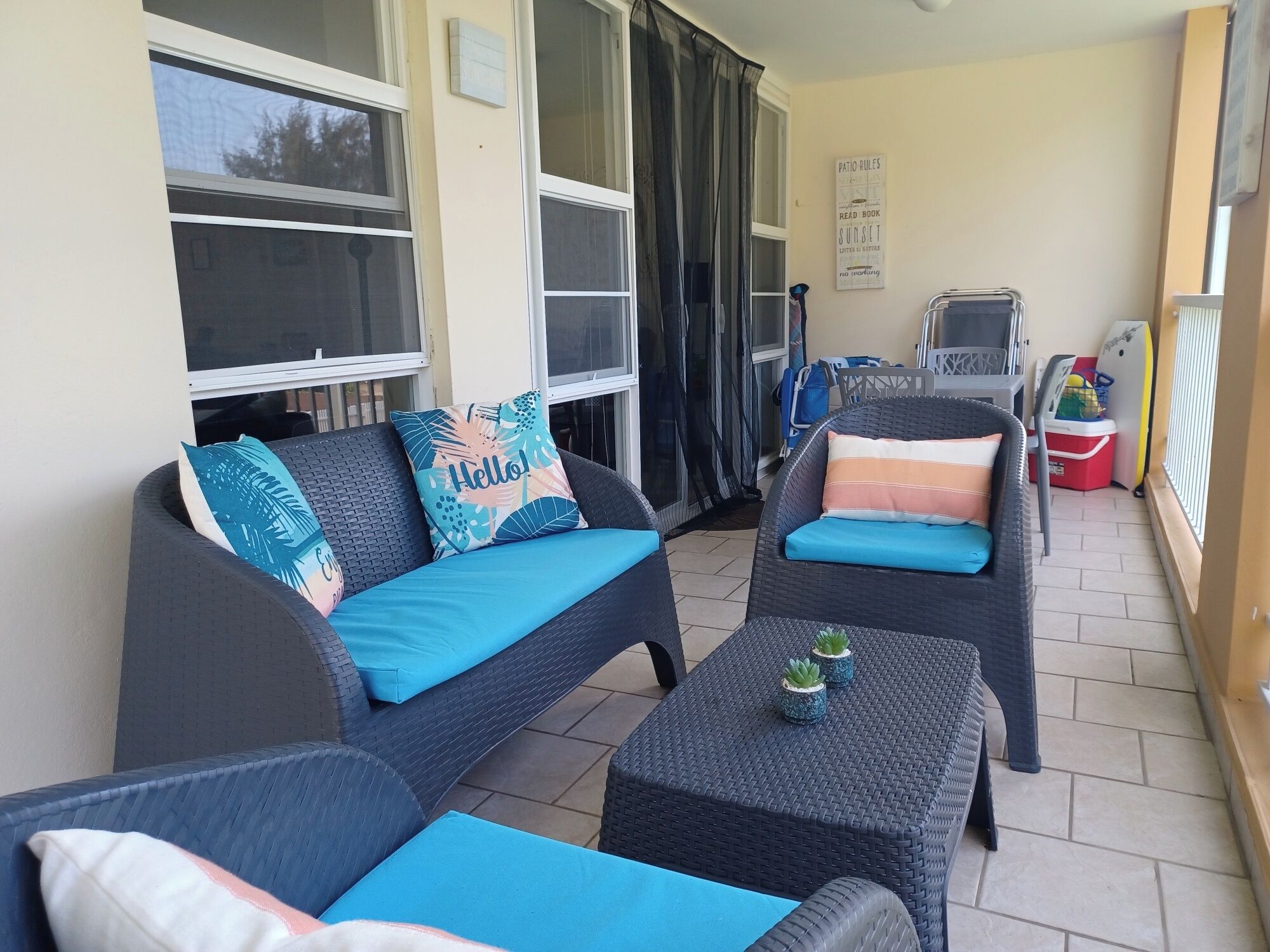 Un patio con mobiliario de mimbre, que incluye un sofá y dos sillones, cada uno con cojines azules. Hay plantas en macetas sobre la mesa, y un cartel en la pared dice 'Patio Rules Read Book Sunset'.