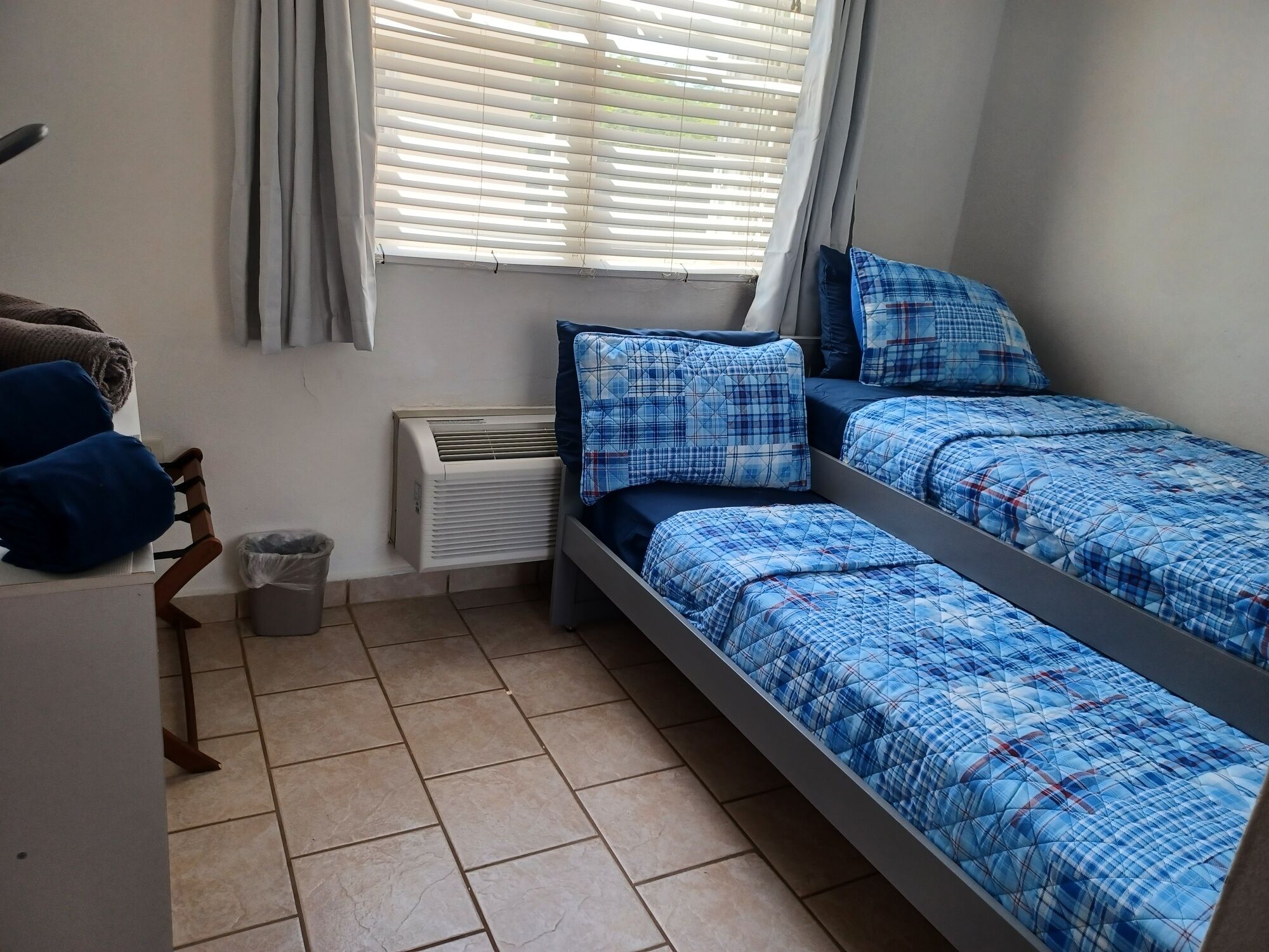 A bedroom with two beds, a window with blinds, an air conditioner, and a tiled floor.