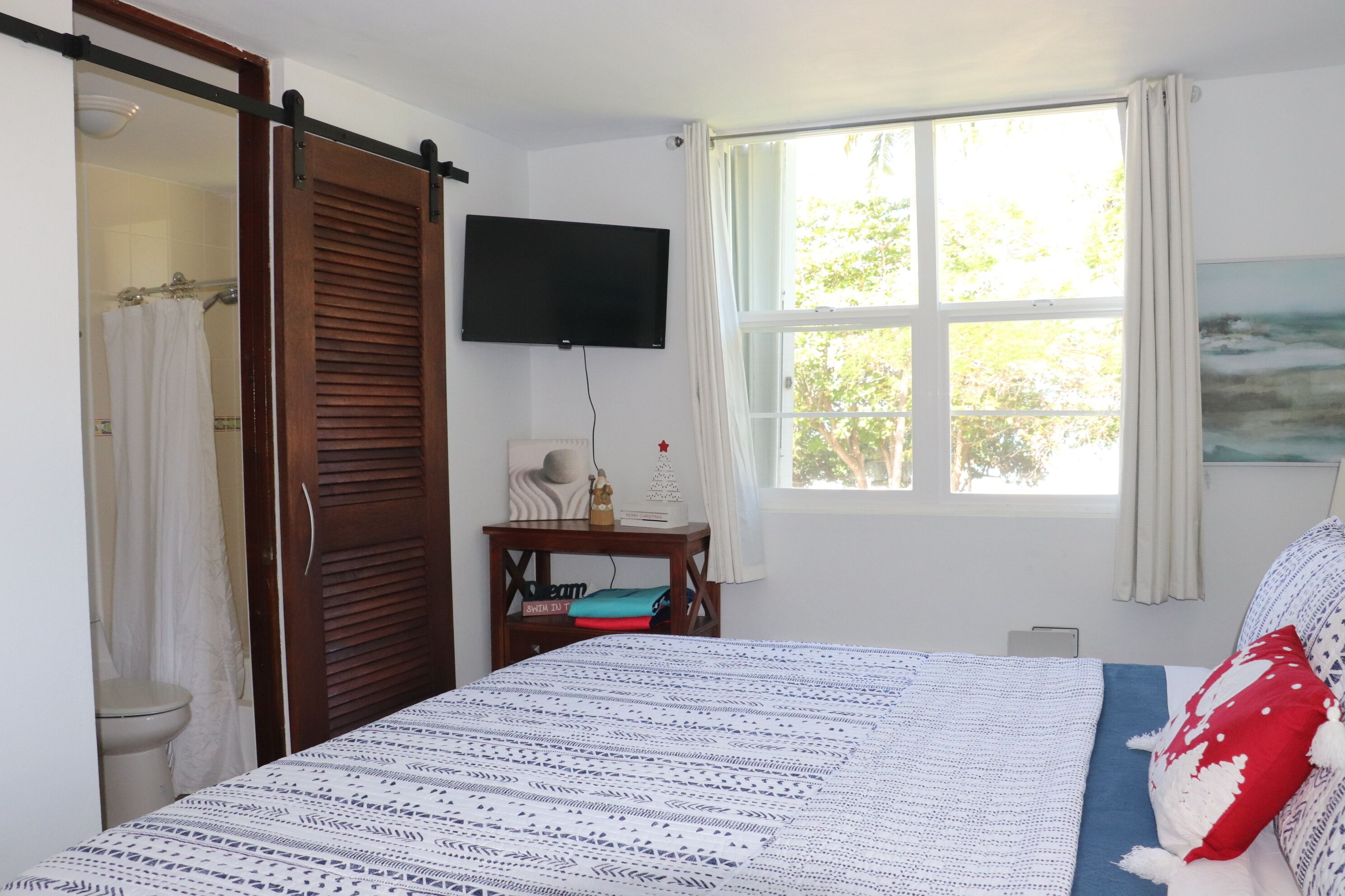A bedroom with a bed, a TV mounted on the wall, a window with curtains, and a painting on the wall.
